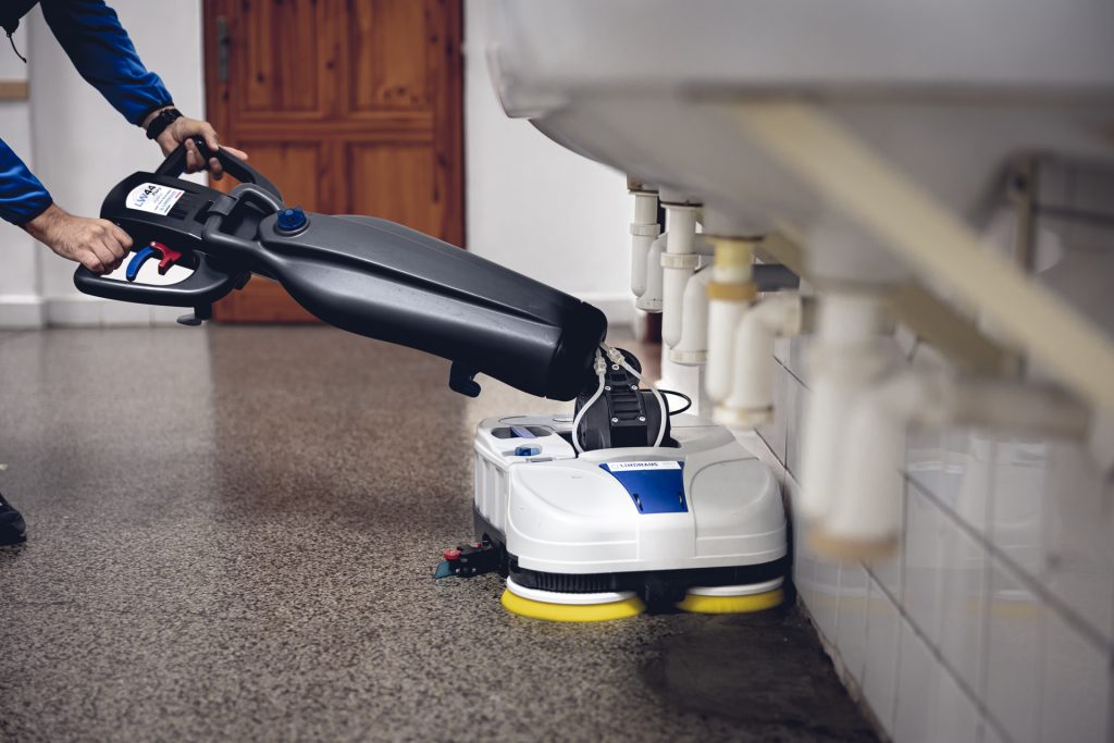 Muž čistí liatu podlahu pod umývadlami v školskej kúpeľni pomocou podlahového umývacieho stroja LINDHAUS LW44 Flexy